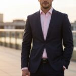 Mężczyzna w granatowym garniturze i różowej koszuli na tle rozmytego miejskiego krajobrazu o zachodzie słońca