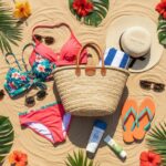 Letni zestaw plażowy na piasku: stroje kąpielowe, okulary, kapelusz, ręcznik, krem, klapki i torba.